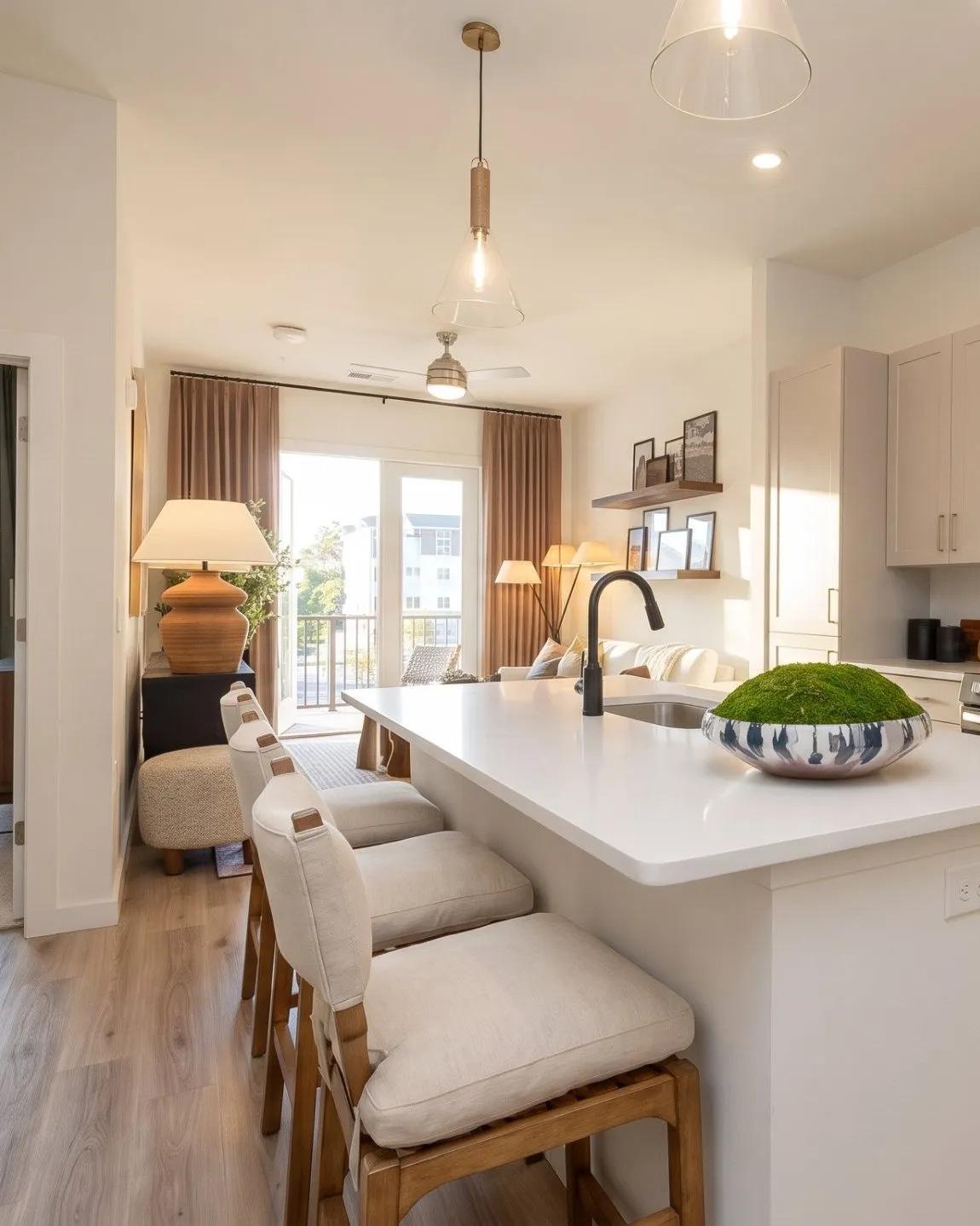 A sunlit kitchen with a white quartz island and linen counter stools, pale shaker cabinets, and the living room and bedroom visible through the doorway beyond